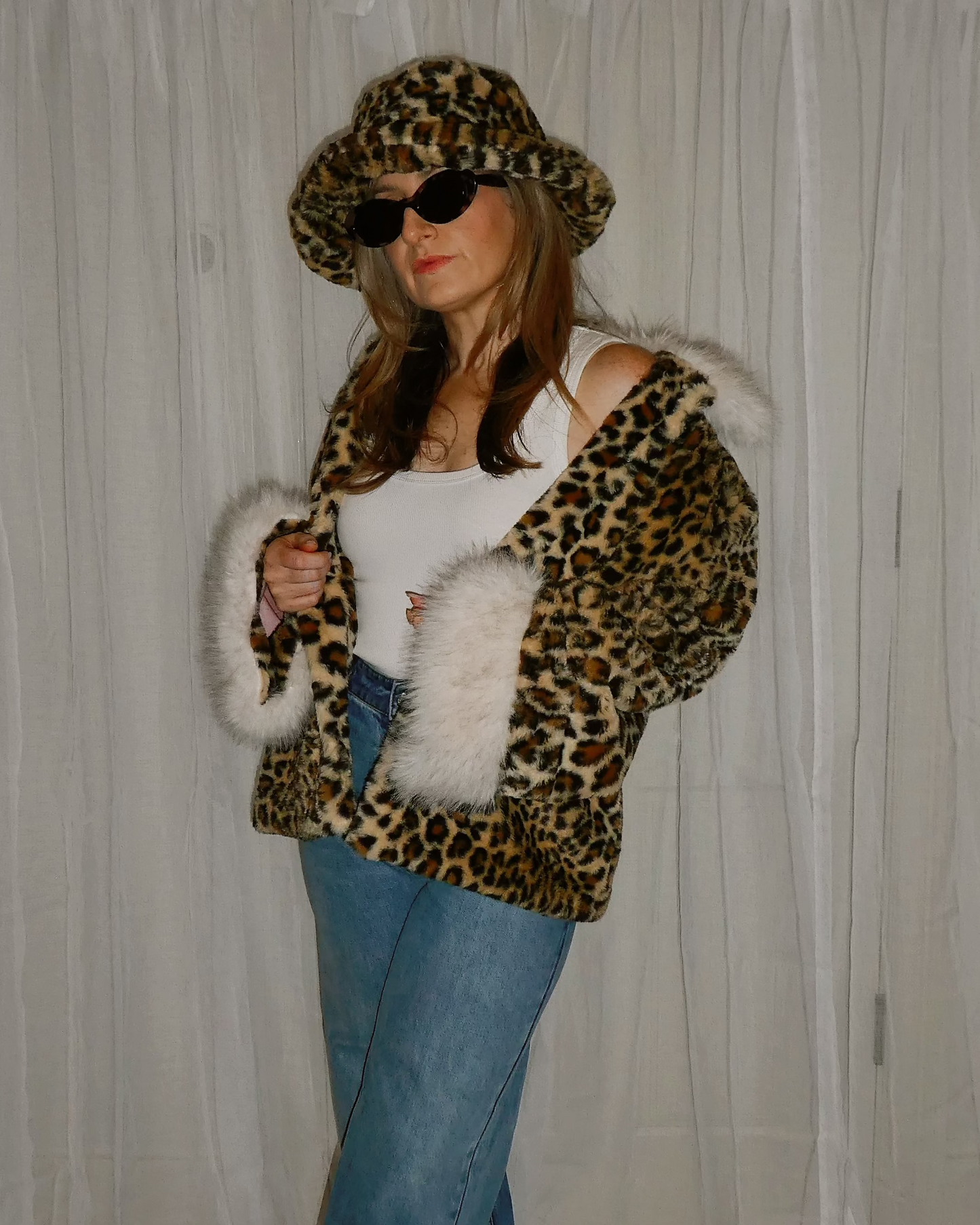 Person wearing a leopard print jacket with a hat sunglasses, and blue jeans in front of a white curtain.