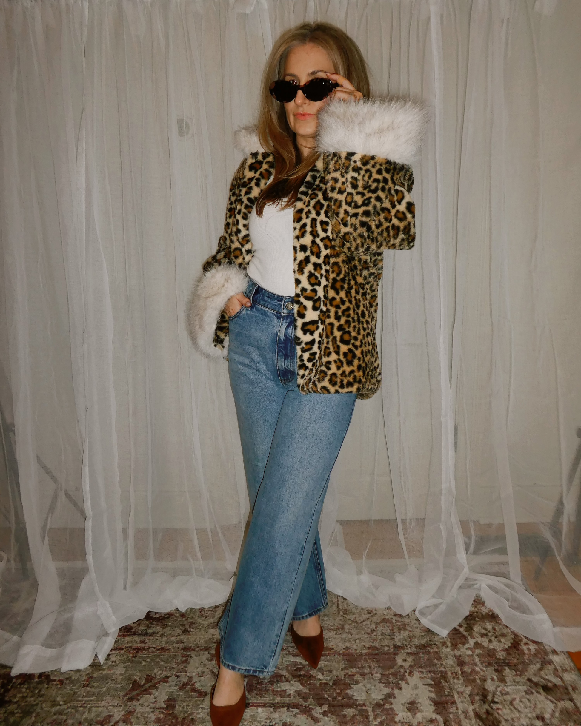 Person wearing a leopard print jacket with fur trim, white top, and blue jeans in front of sheer curtains.