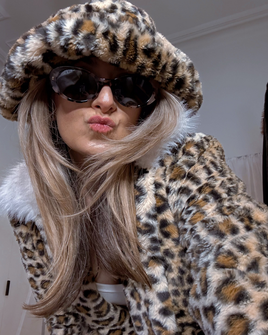 Person wearing a leopard print coat and hat with sunglasses, blowing a kiss.