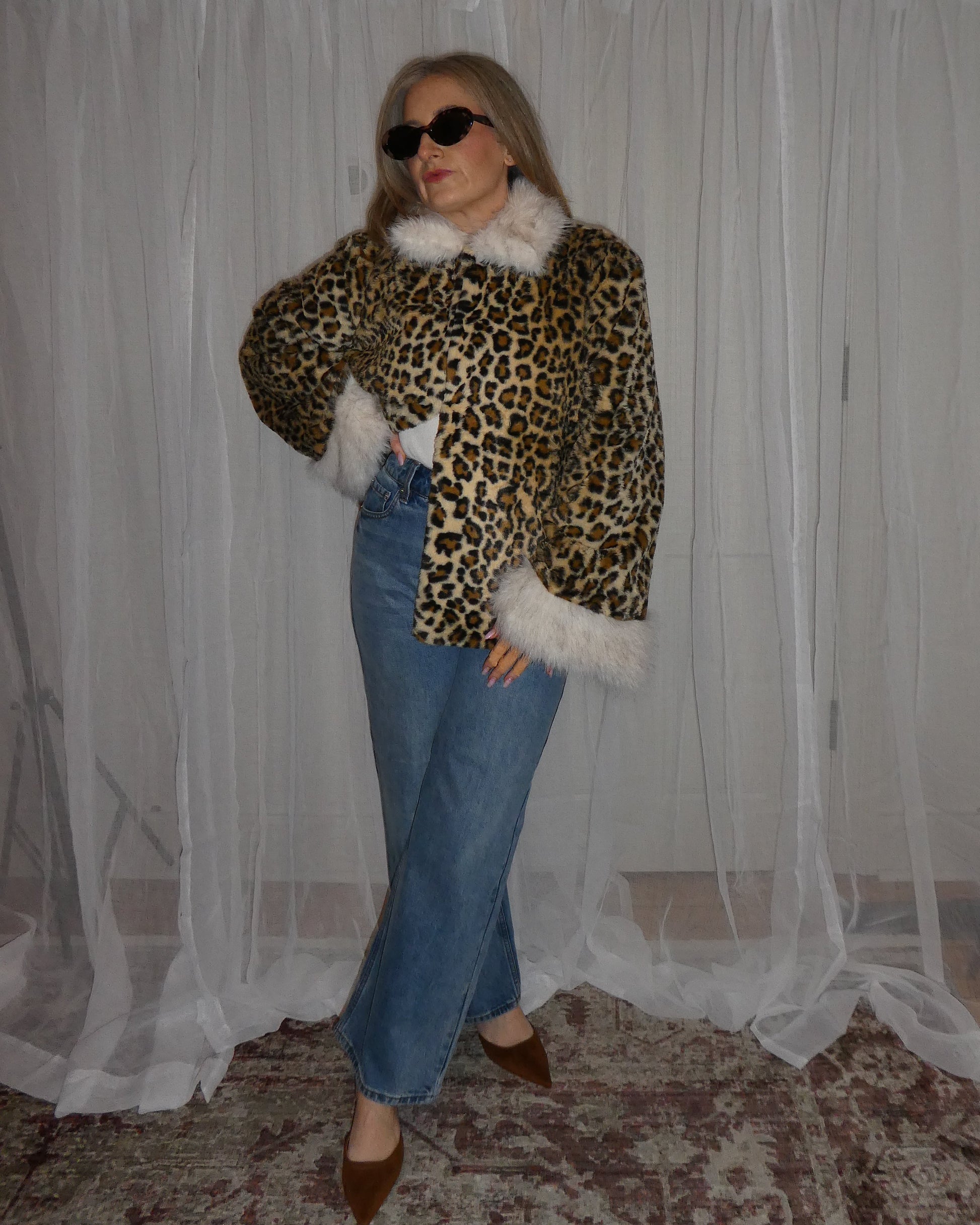 Person wearing a leopard print jacket with white fur trim in front of a white curtain.