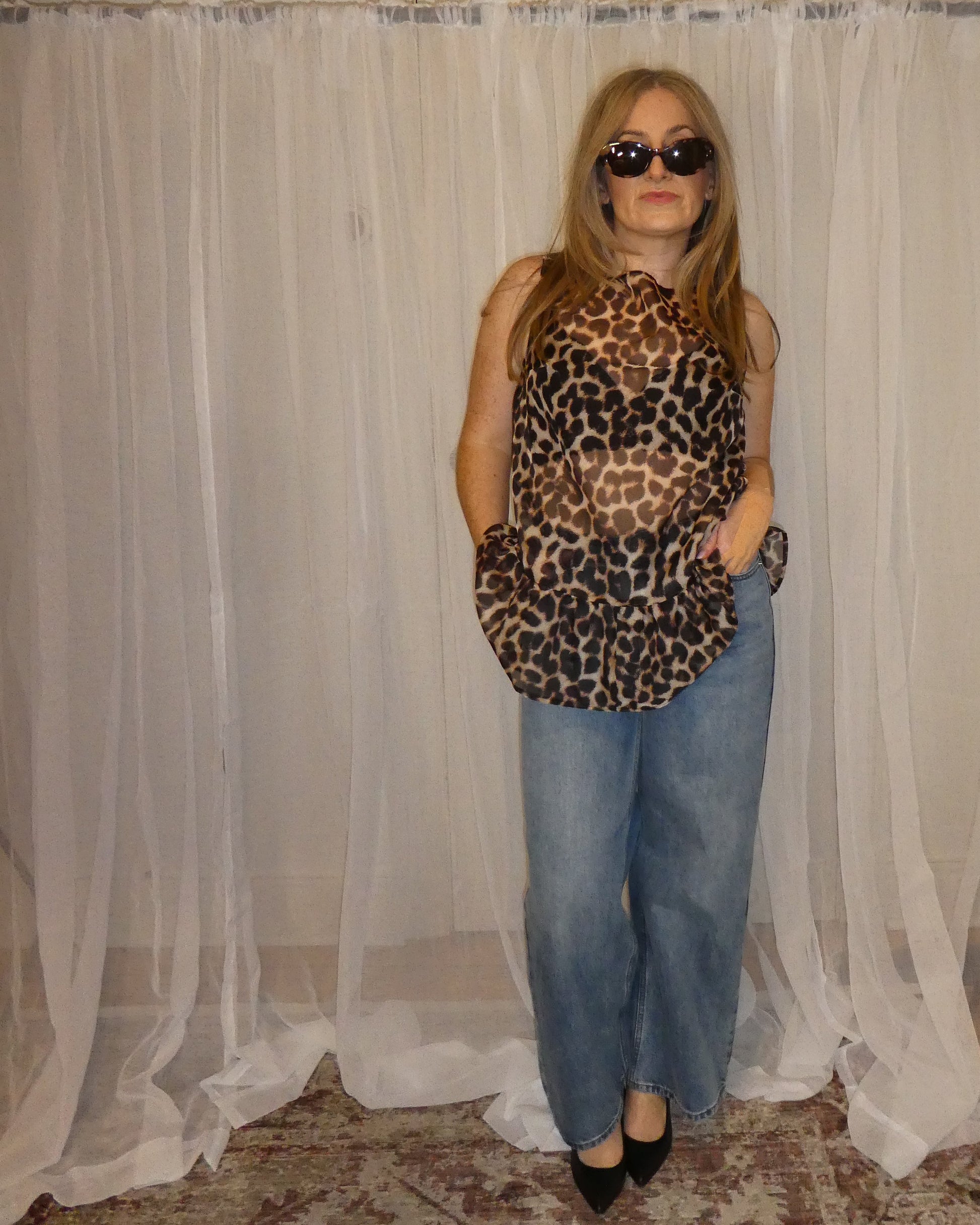 Woman wearing a leopard print top and blue jeans standing in front of a white curtain.