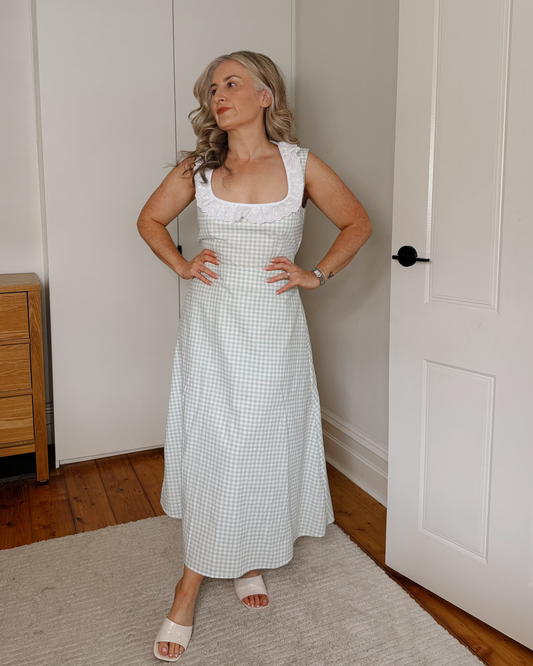 A woman stands in a bedroom wearing a white sleeveless midi dress with a small light blue gingham pattern. She is standing on a light-colored rug in front of white wardrobes and a wooden dresser.