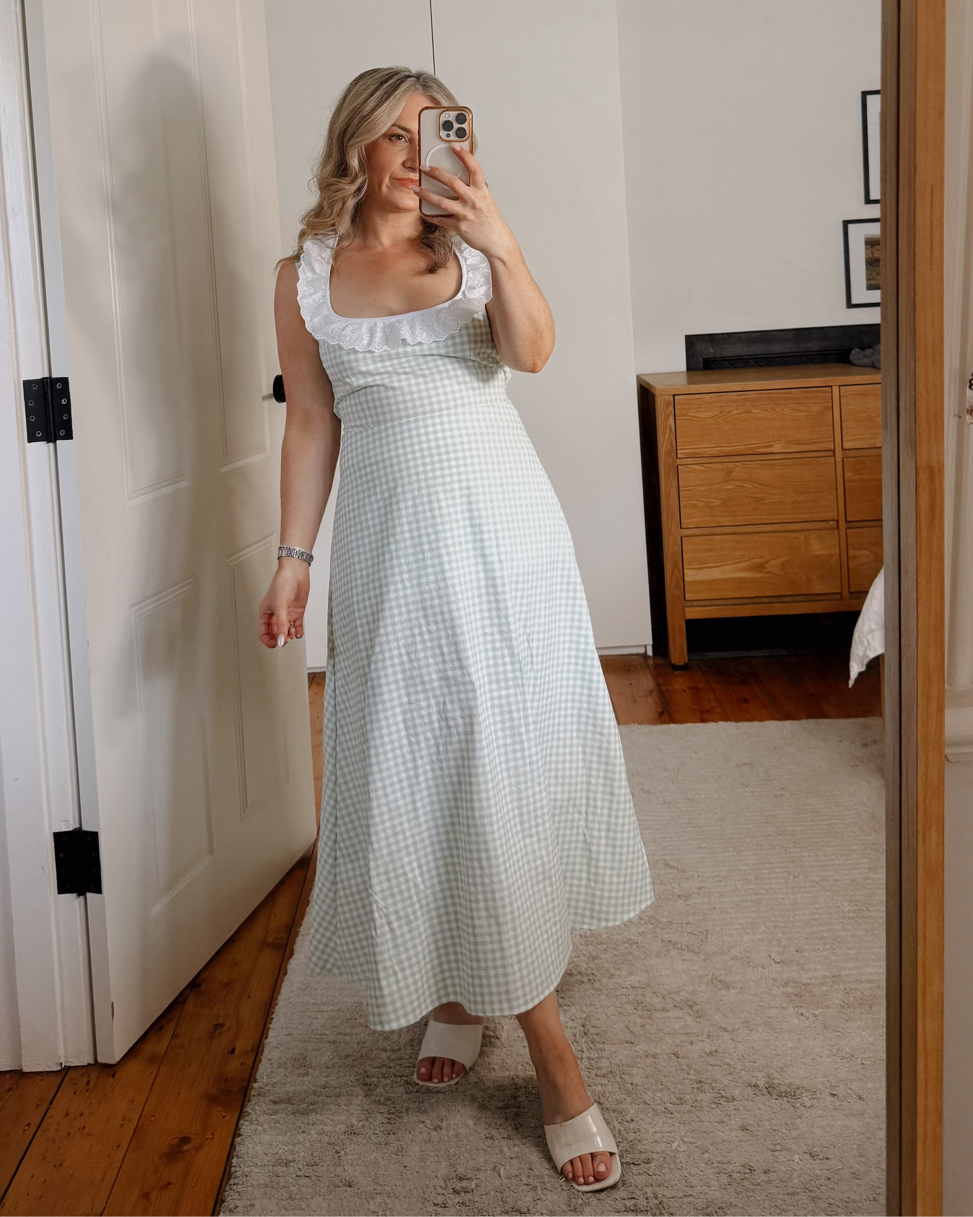 A woman takes a mirror selfie in a home setting, wearing a white sleeveless midi dress with a subtle gingham pattern and white slide sandals.