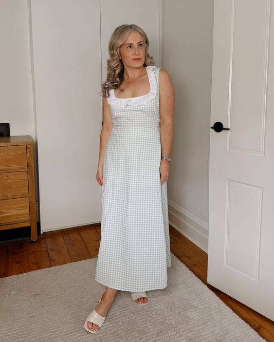 A woman wearing a white and light green gingham maxi dress with a white lace-trimmed scoop neckline, standing in a bedroom with white walls, a wooden dresser, and a light-colored rug.