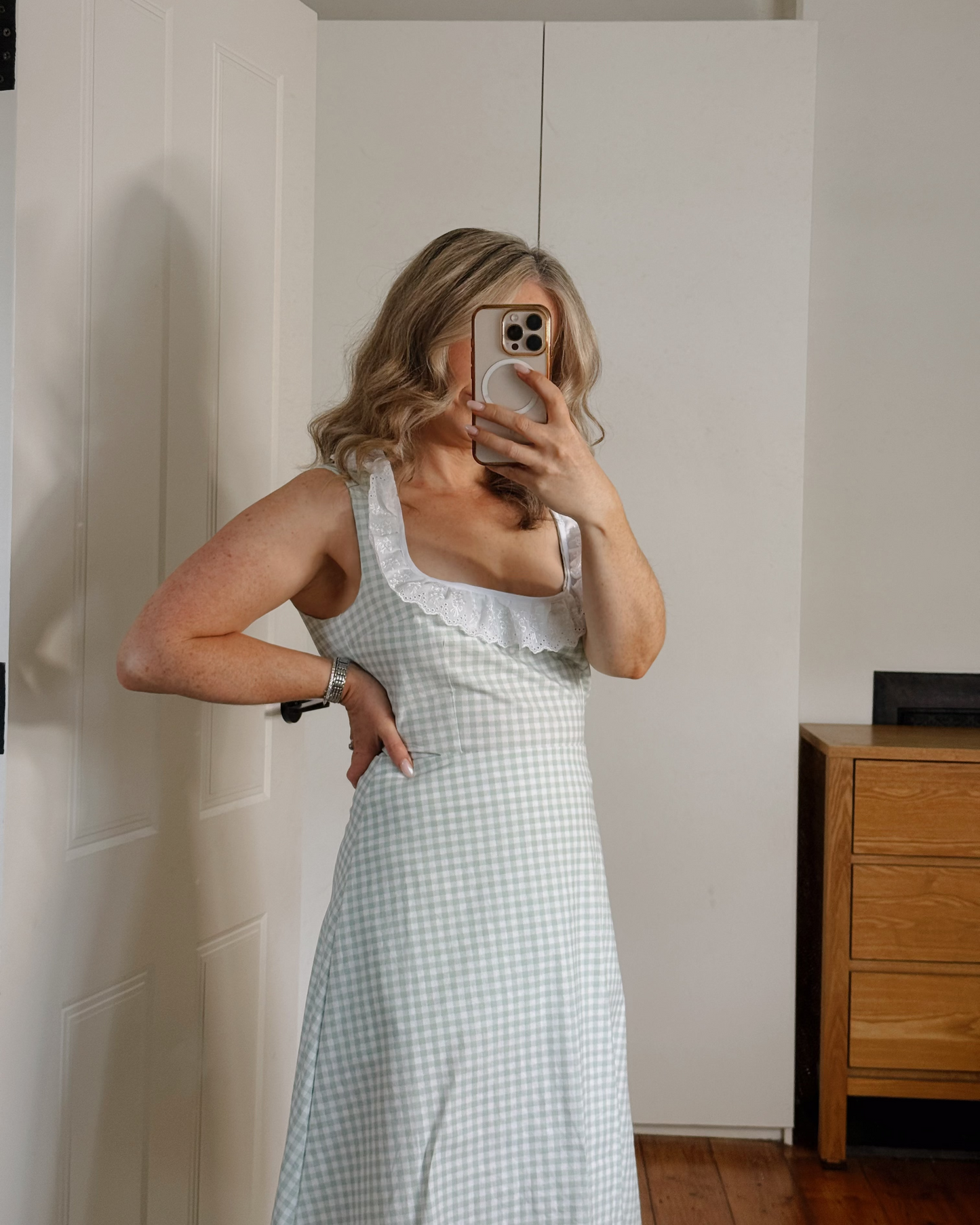 A woman takes a mirror selfie wearing a light blue and white gingham pinafore dress with a white ruffled neckline, standing in a room with white walls and a wooden dresser.