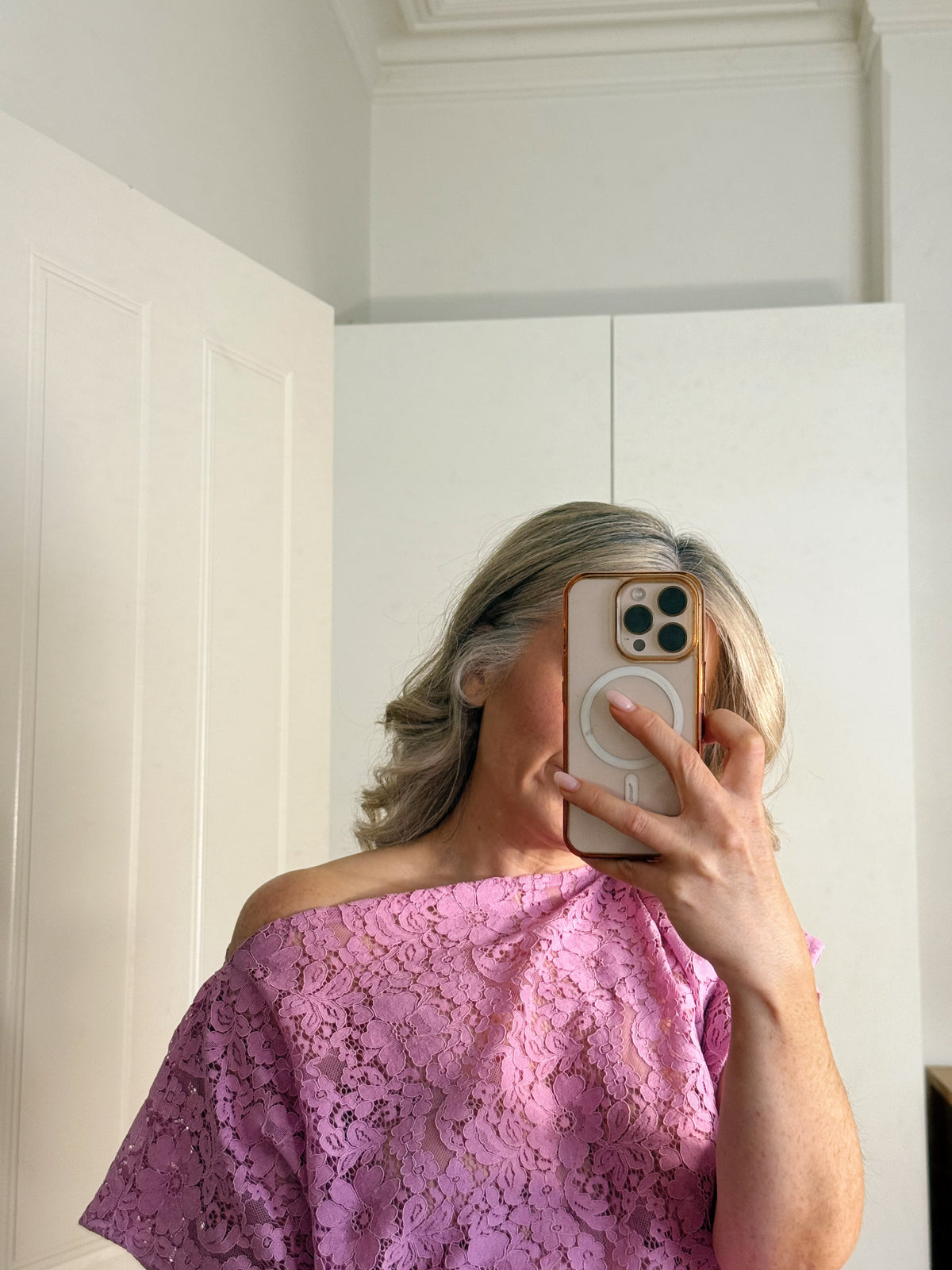 A woman taking a mirror selfie wearing a pink off-the-shoulder lace top, holding a phone with a MagSafe case in a bright room with white walls and doors.