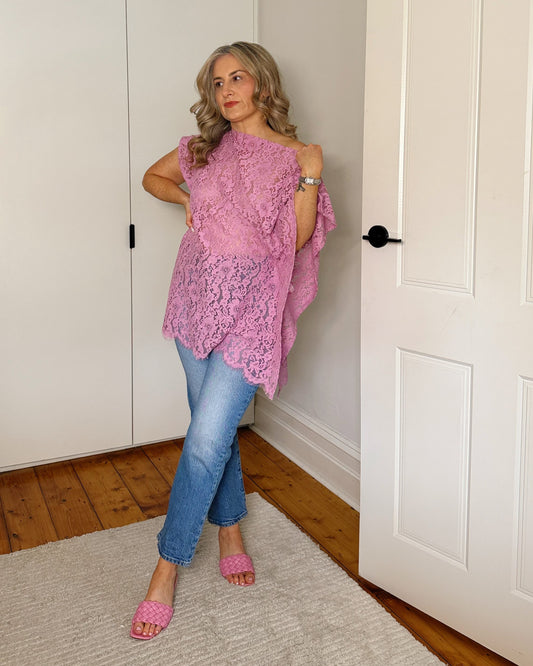 A woman modeling a pink lace top and matching pink flat sandals, paired with blue jeans, standing in a room with white walls, a white wardrobe, and a wooden floor.