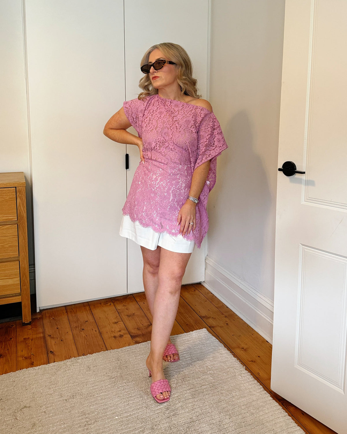 A woman modeling a pink lace off-the-shoulder tunic top with a white underlayer and pink knit slide sandals, standing in a room with white doors and wooden floors.