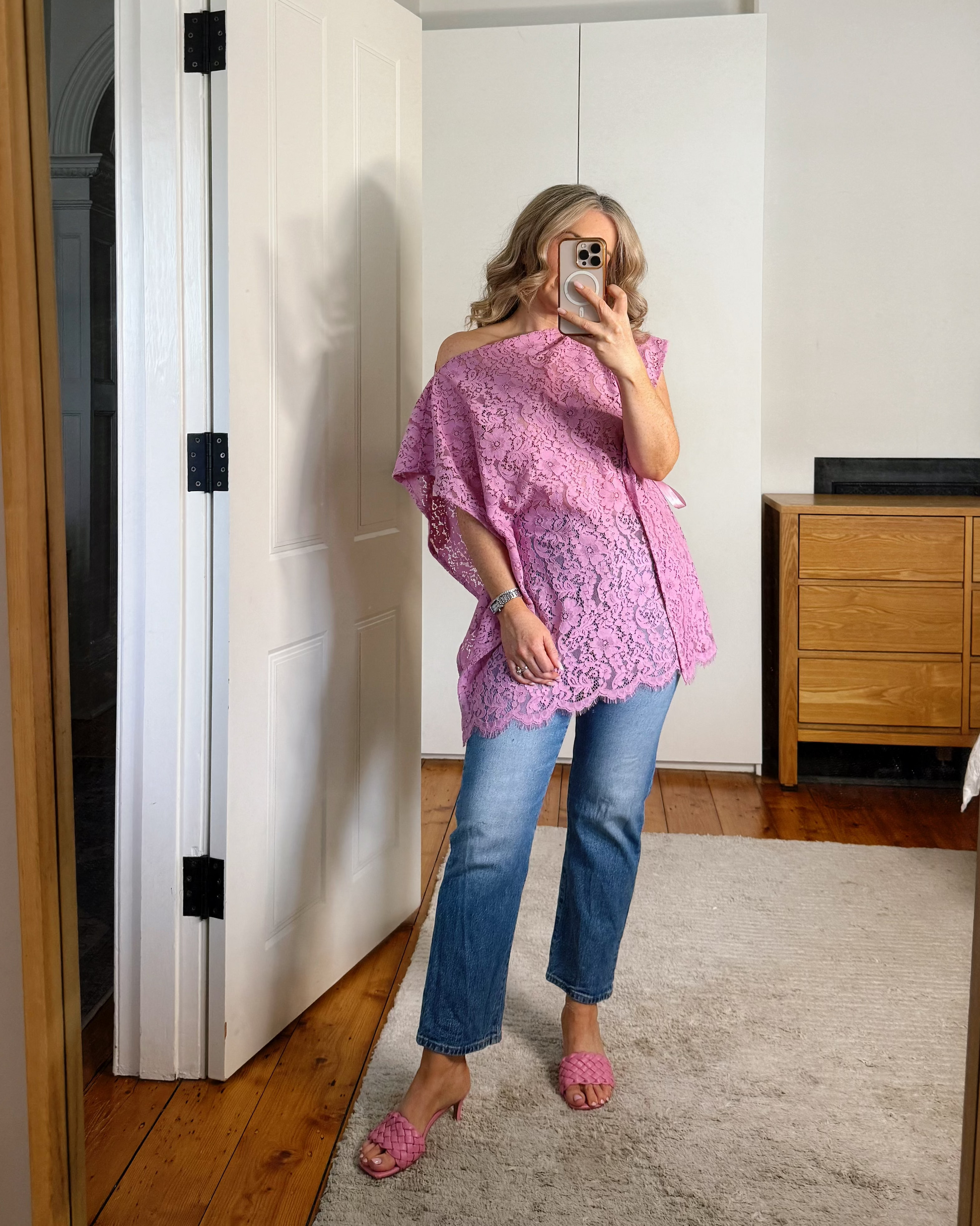 A woman takes a mirror selfie in a room, wearing a pink off-the-shoulder lace top, blue jeans, and pink flat shoes.