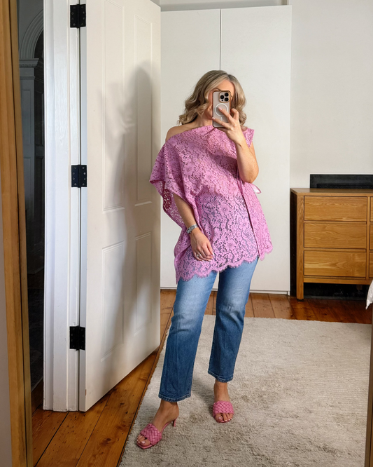 A woman takes a mirror selfie in a room, wearing a pink off-the-shoulder lace top, blue jeans, and pink flat shoes.