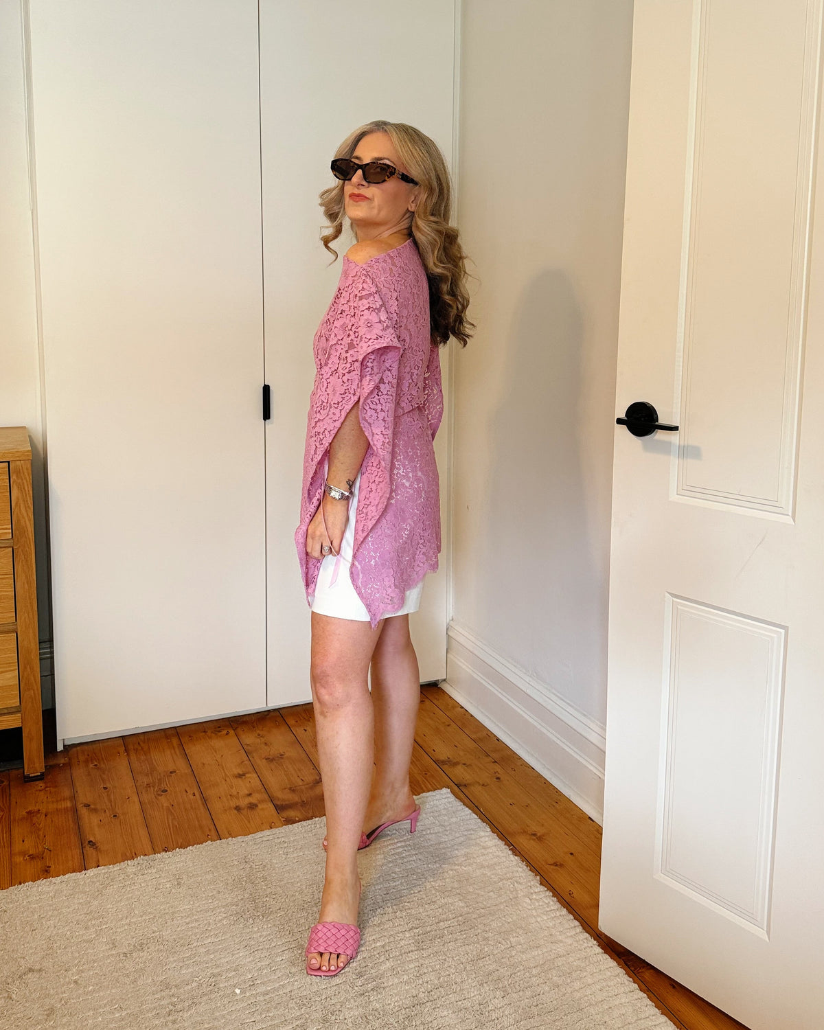 A woman wearing a pink open-front knit poncho-style top, white shorts, and pink flat sandals stands in a room with white walls, a white wardrobe, a wooden dresser, and a light rug.