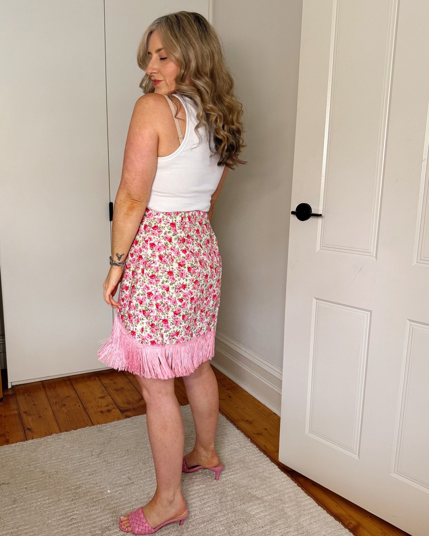 A woman stands in a bedroom wearing a white sleeveless top and a pink floral fringed skirt, paired with pink flat shoes.