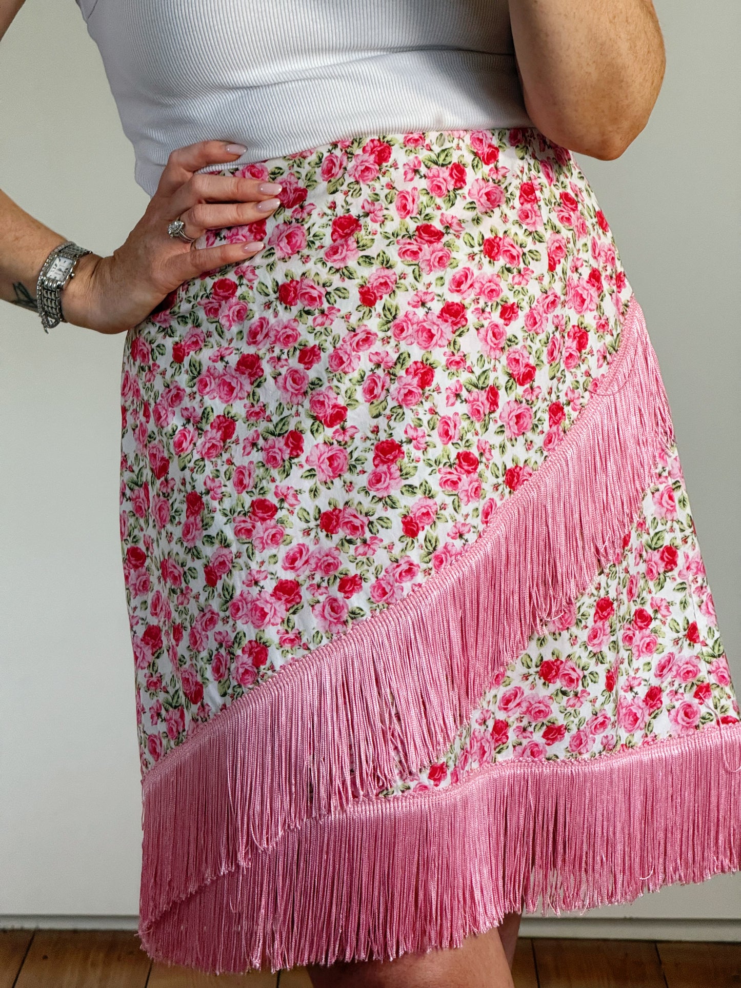 A close-up of a person wearing a pink and red floral skirt with a diagonal fringe panel and a long bottom fringe hem, paired with a white ribbed top.