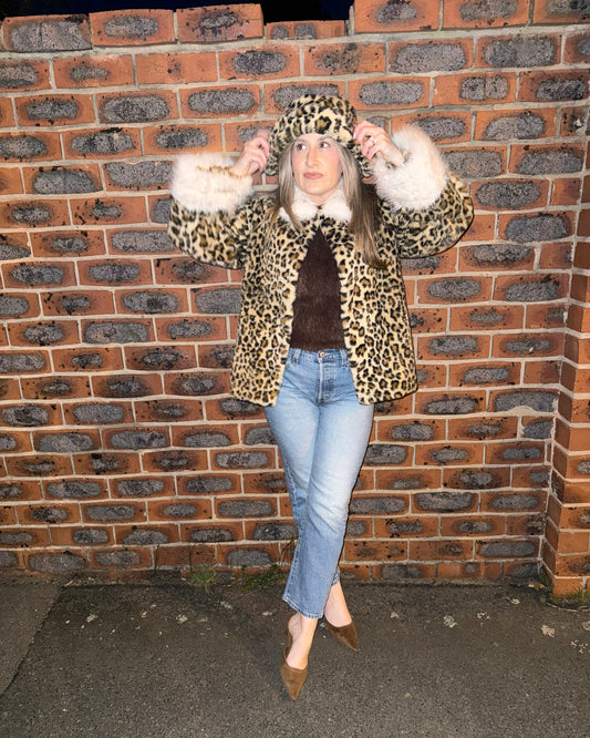 A woman wearing a leopard print faux fur coat with white fur trim, a brown top, light blue jeans, and brown pointed-toe flats, posing in front of a red brick wall.