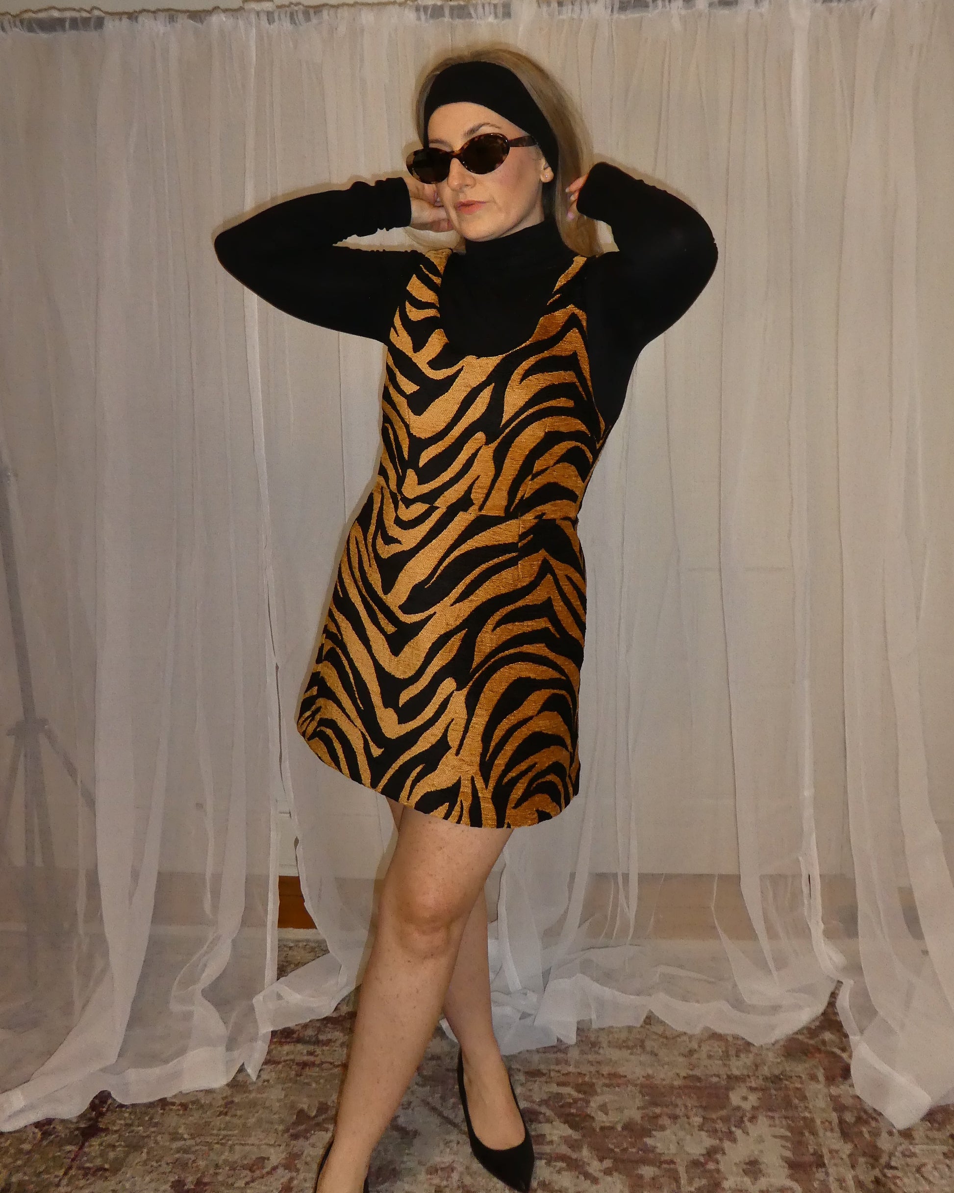 Person wearing a black and orange tiger-striped dress standing in front of sheer curtains.