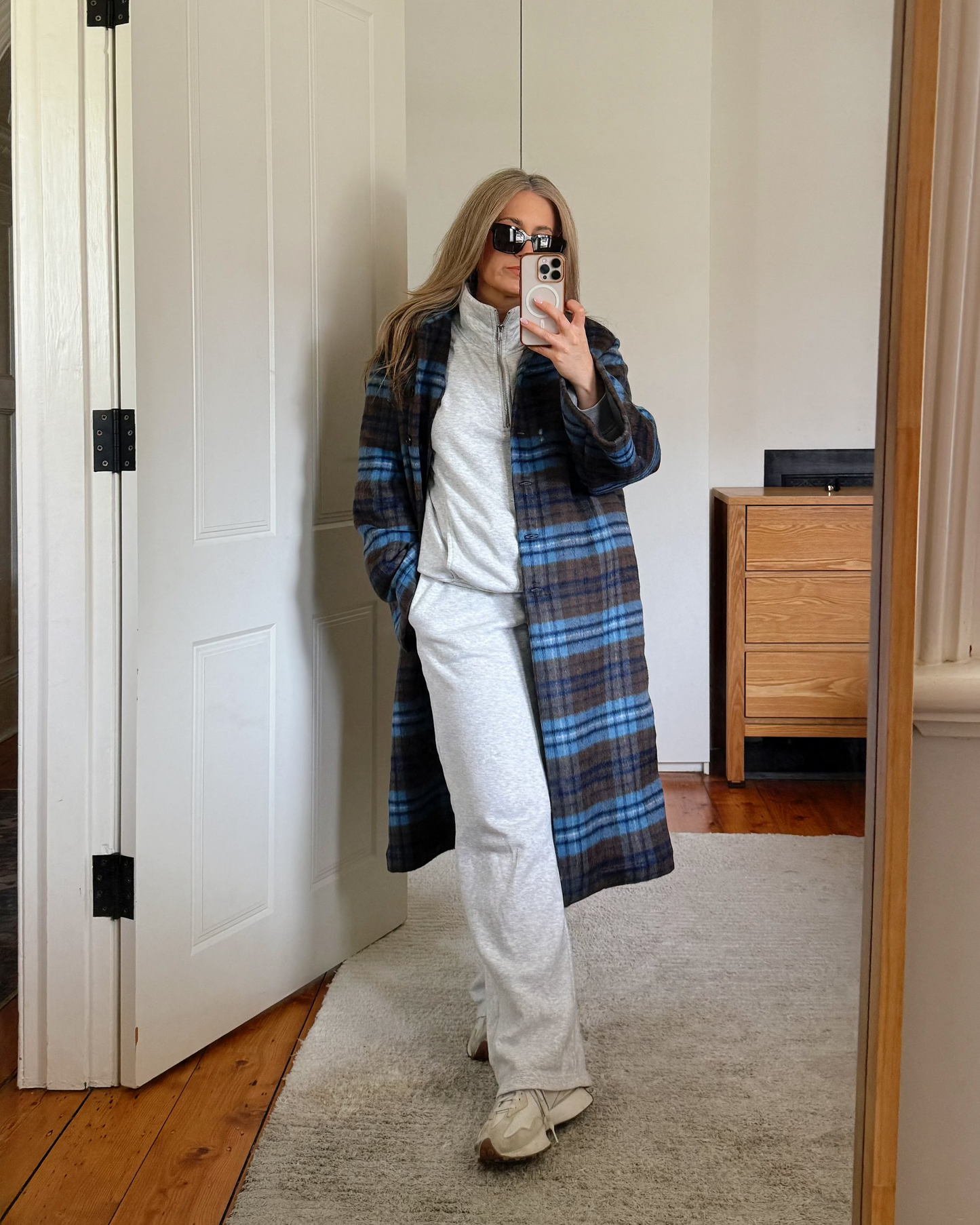 A woman takes a mirror selfie in a room, wearing a long blue and brown plaid coat over a grey matching track suit.