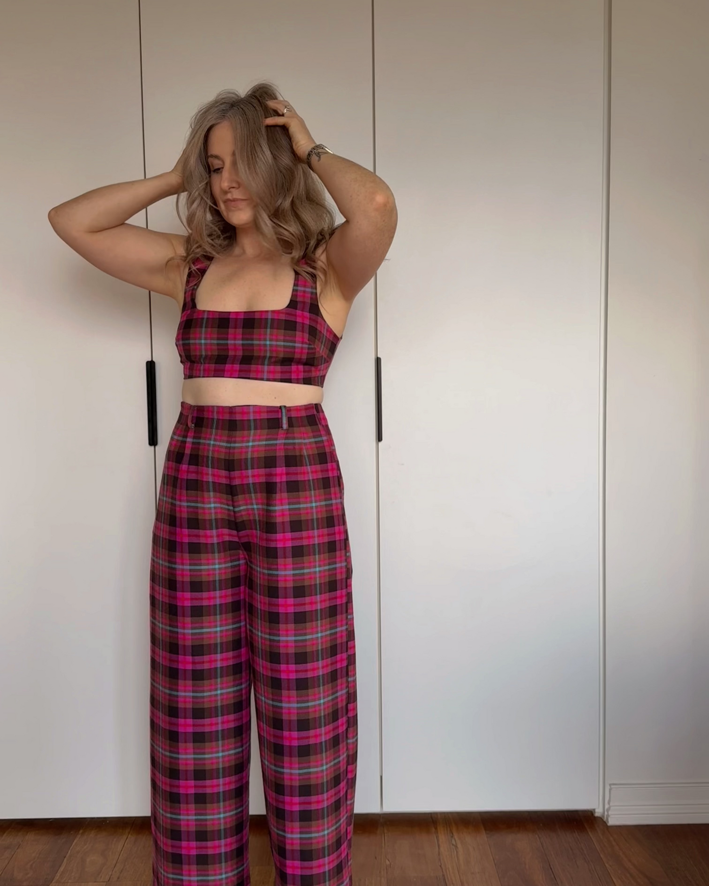 A woman stands indoors against a white wardrobe door wearing a matching plaid crop top and wide-leg pants set in bright pink, blue and brown tartan.