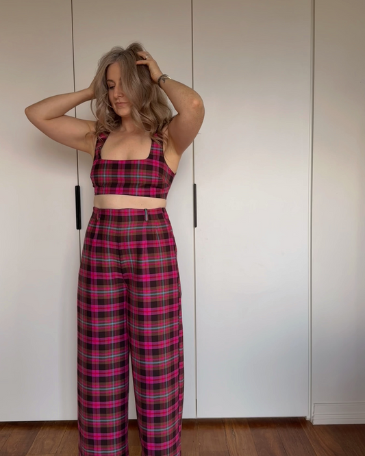 A woman stands indoors against a white wardrobe door wearing a matching plaid crop top and wide-leg pants set in bright pink, blue and brown tartan.