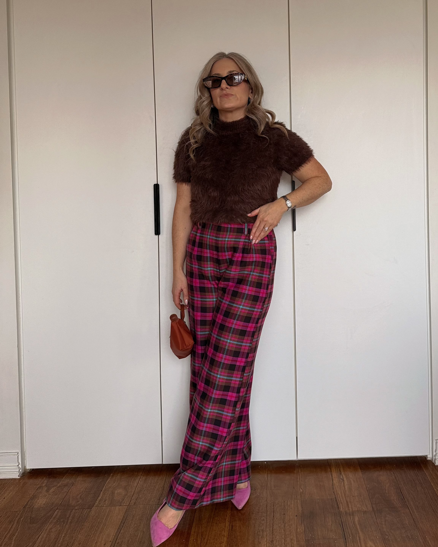 A woman posing in front of white closet doors wearing a brown fuzzy short-sleeve top, pink and red plaid trousers, and pink pointed shoes, holding a small brown handbag.