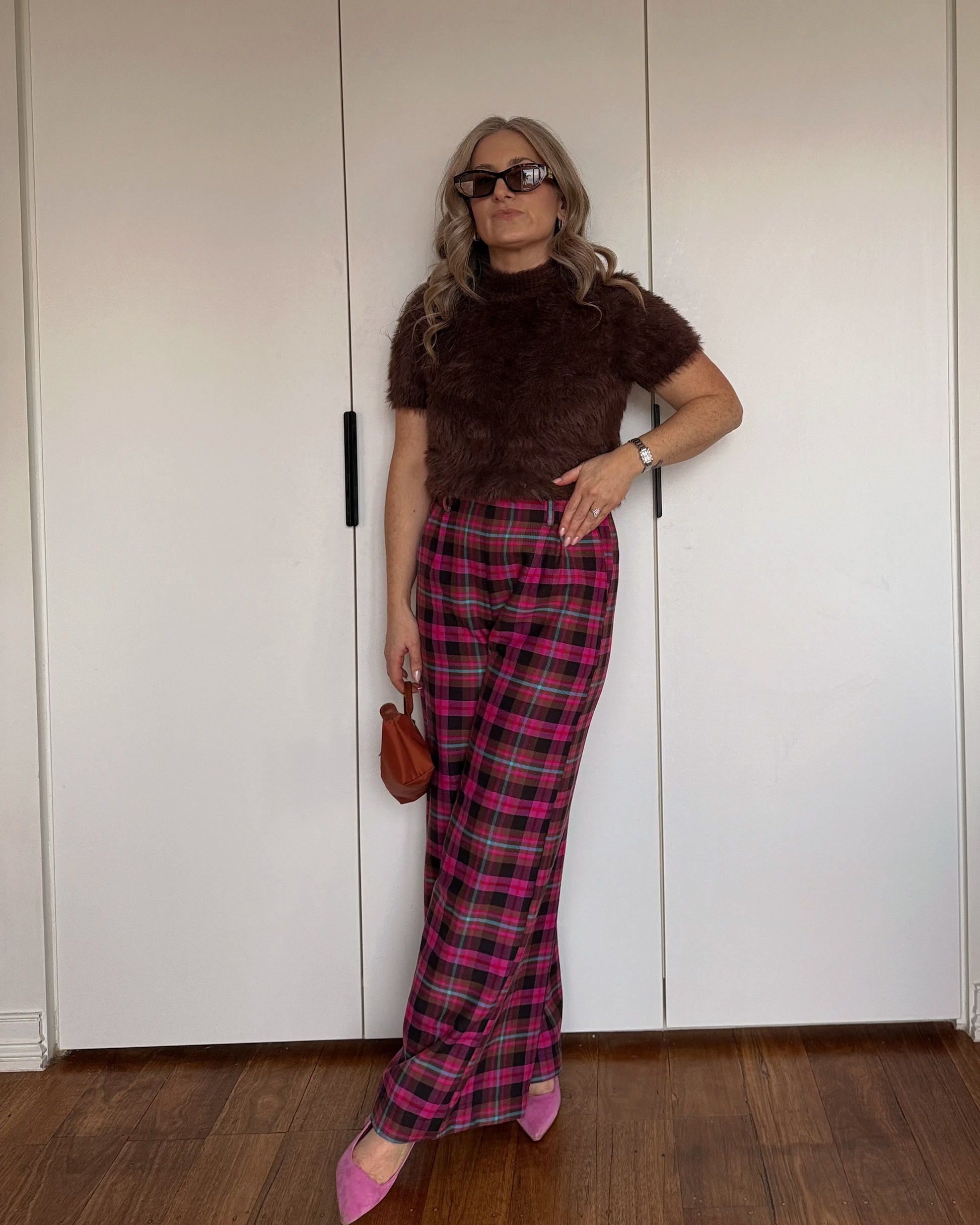 A woman posing in front of white closet doors wearing a brown fuzzy short-sleeve top, pink and red plaid trousers, and pink pointed shoes, holding a small brown handbag.