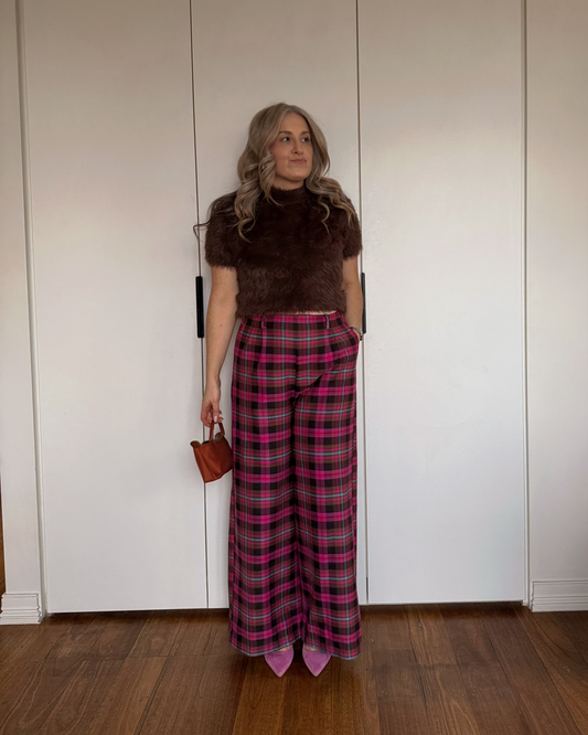 A woman stands in front of white wardrobe doors wearing a dark brown fuzzy short-sleeve top and pink and red plaid wide-leg trousers, holding a small brown handbag.