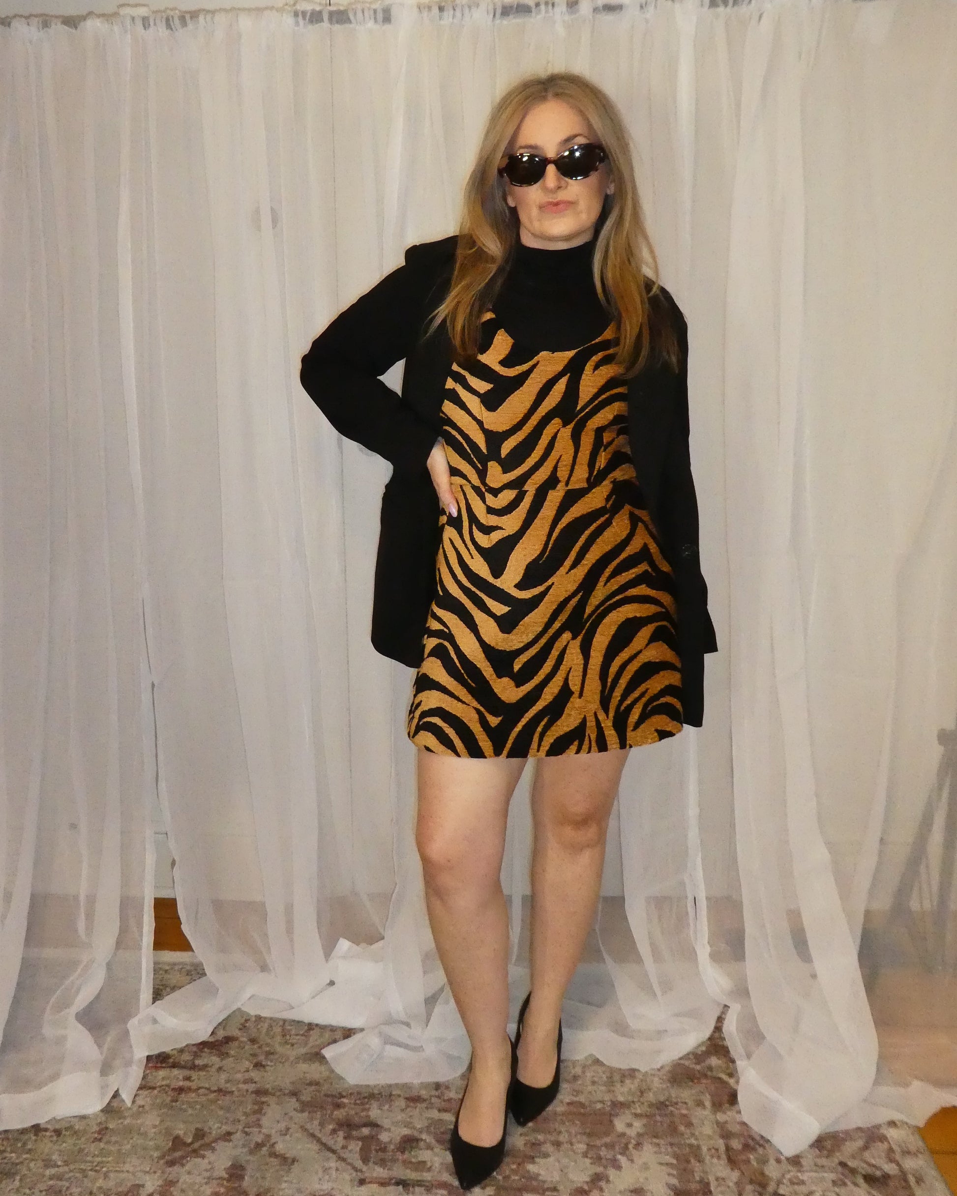 Person wearing a zebra print dress and black coat in front of sheer curtains