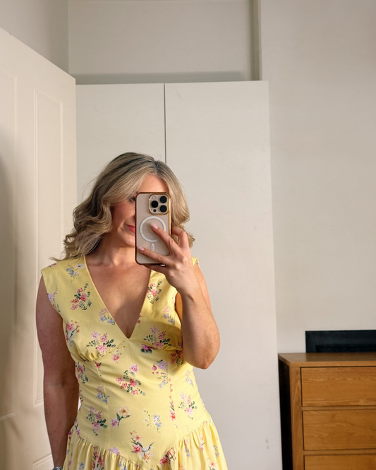 A woman takes a mirror selfie wearing a yellow sleeveless V-neck dress with a pink and green floral pattern. She is standing in a room with white walls, a white wardrobe, and a wooden chest of drawers.