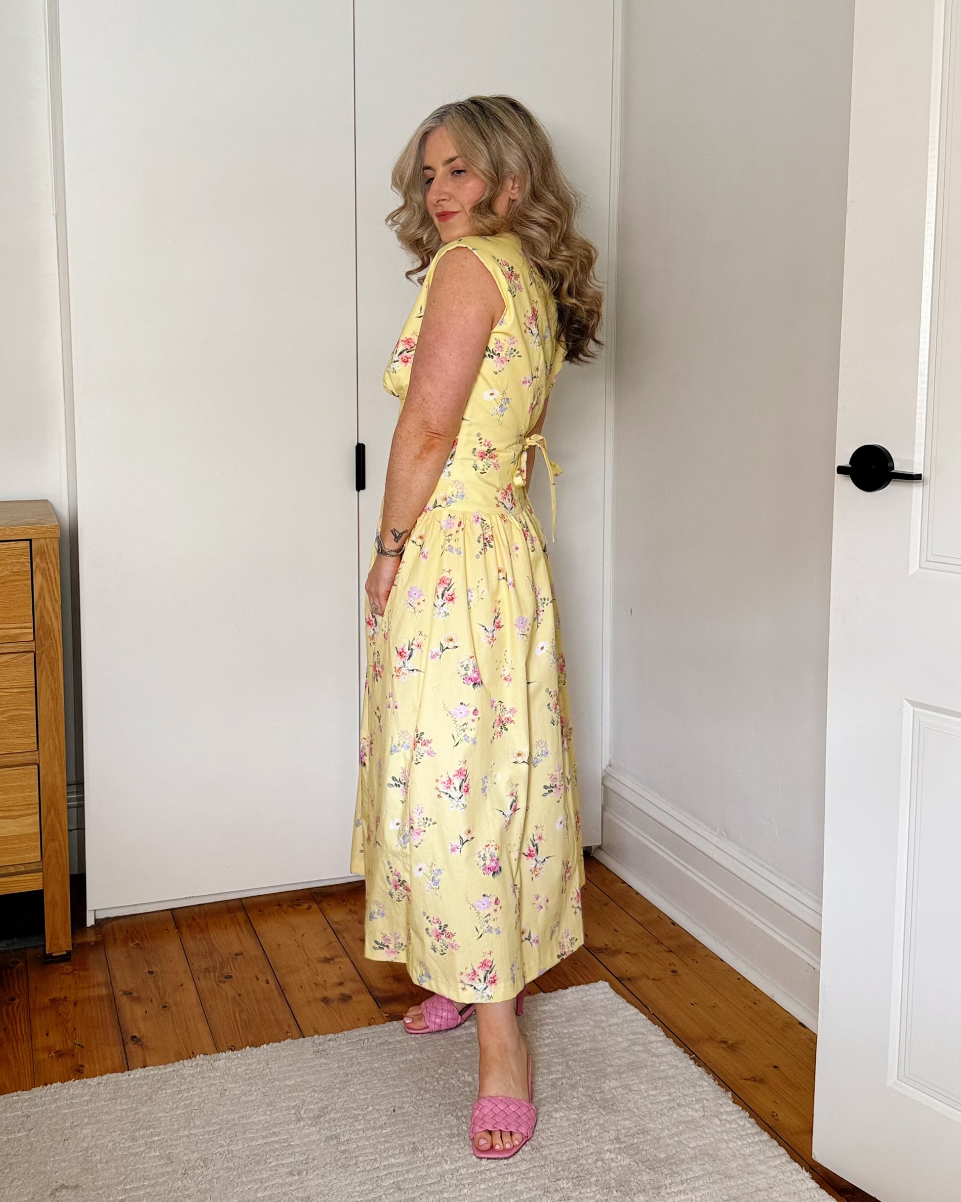 A woman stands in a bedroom wearing a yellow sleeveless floral midi dress with pink shoes, photographed from the side. The room has white walls, a white wardrobe, a wooden dresser, a light rug, and a bed partially visible on the left.