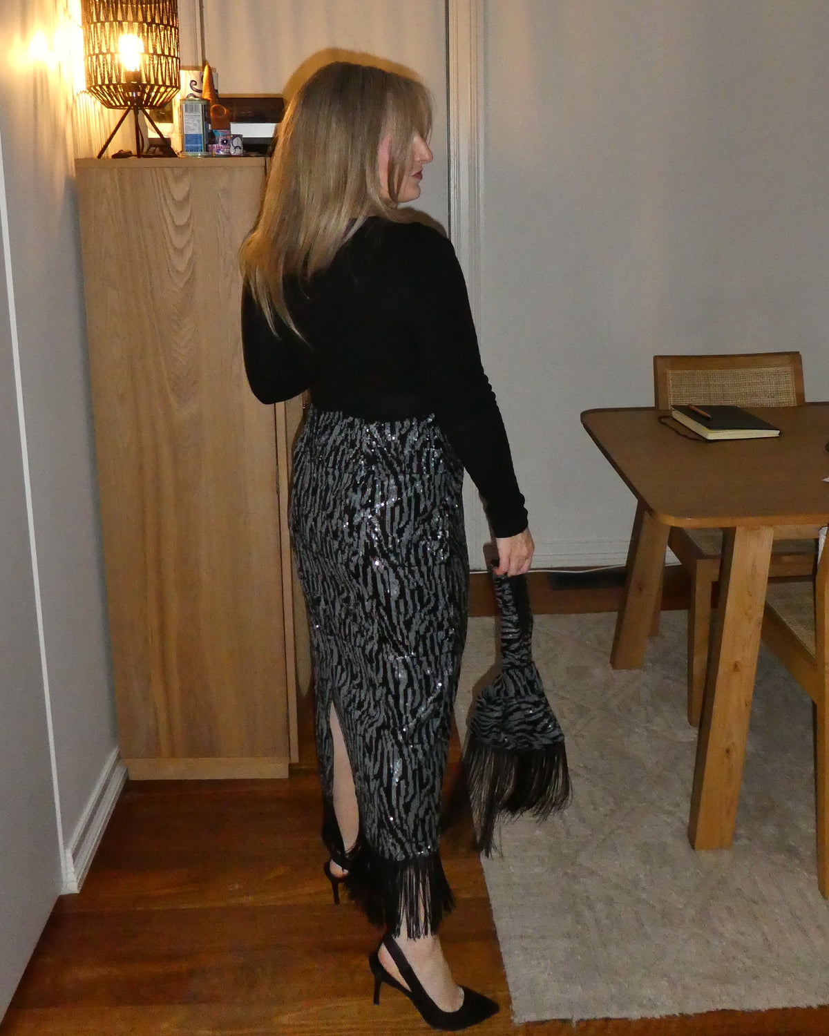 A woman stands in a room wearing a black long-sleeve top and a black-and-white patterned midi skirt with fringe, holding a black fringed accessory. The room has wood flooring, a wooden cabinet, a table, and a lamp.
