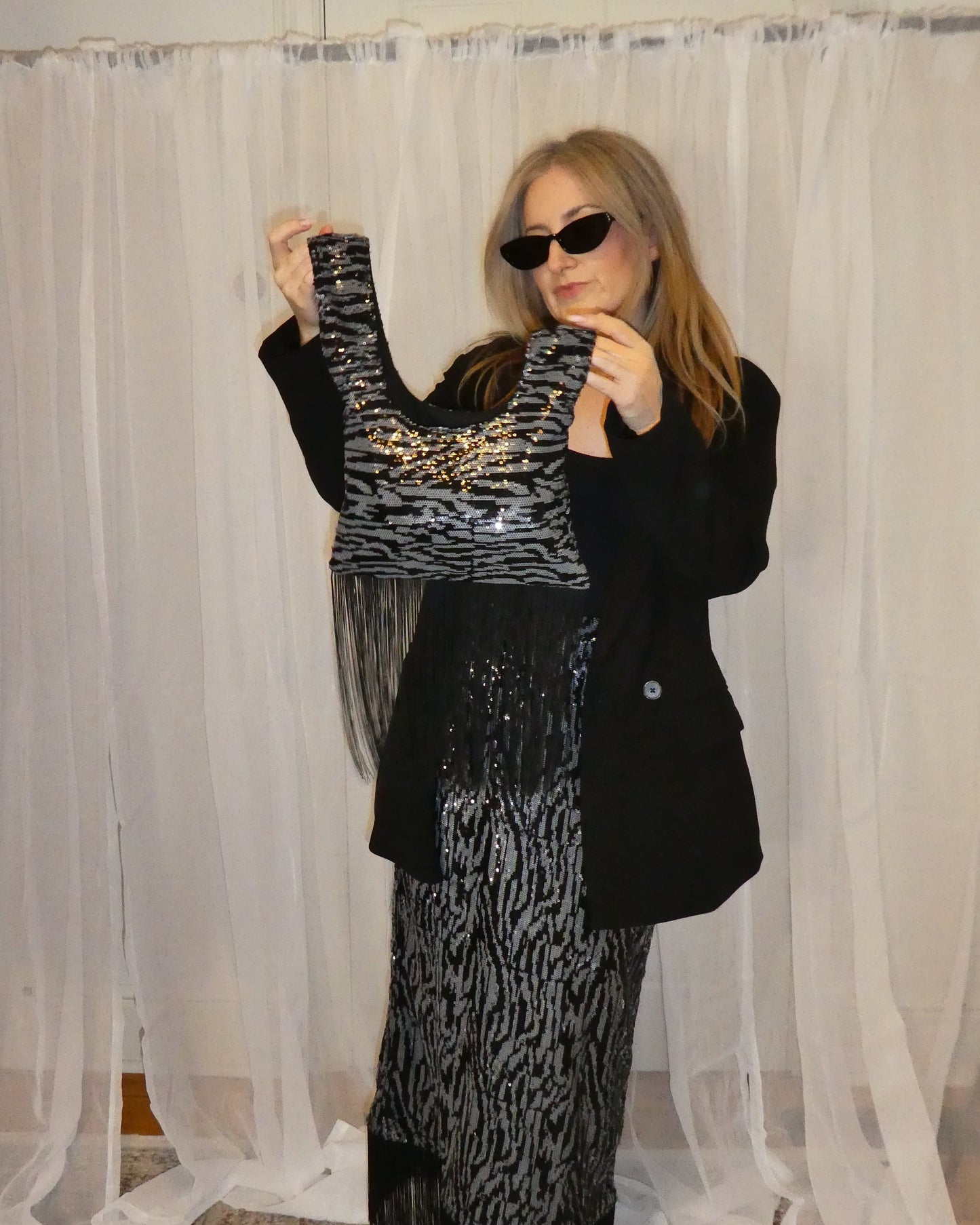 Person wearing a black and silver dress with a black blazer in front of sheer curtains.