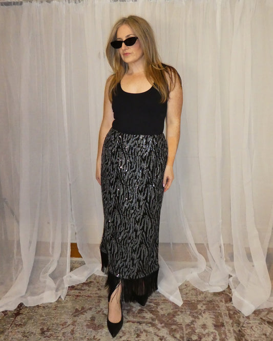 Woman wearing a black top and patterned skirt standing in front of white curtains.