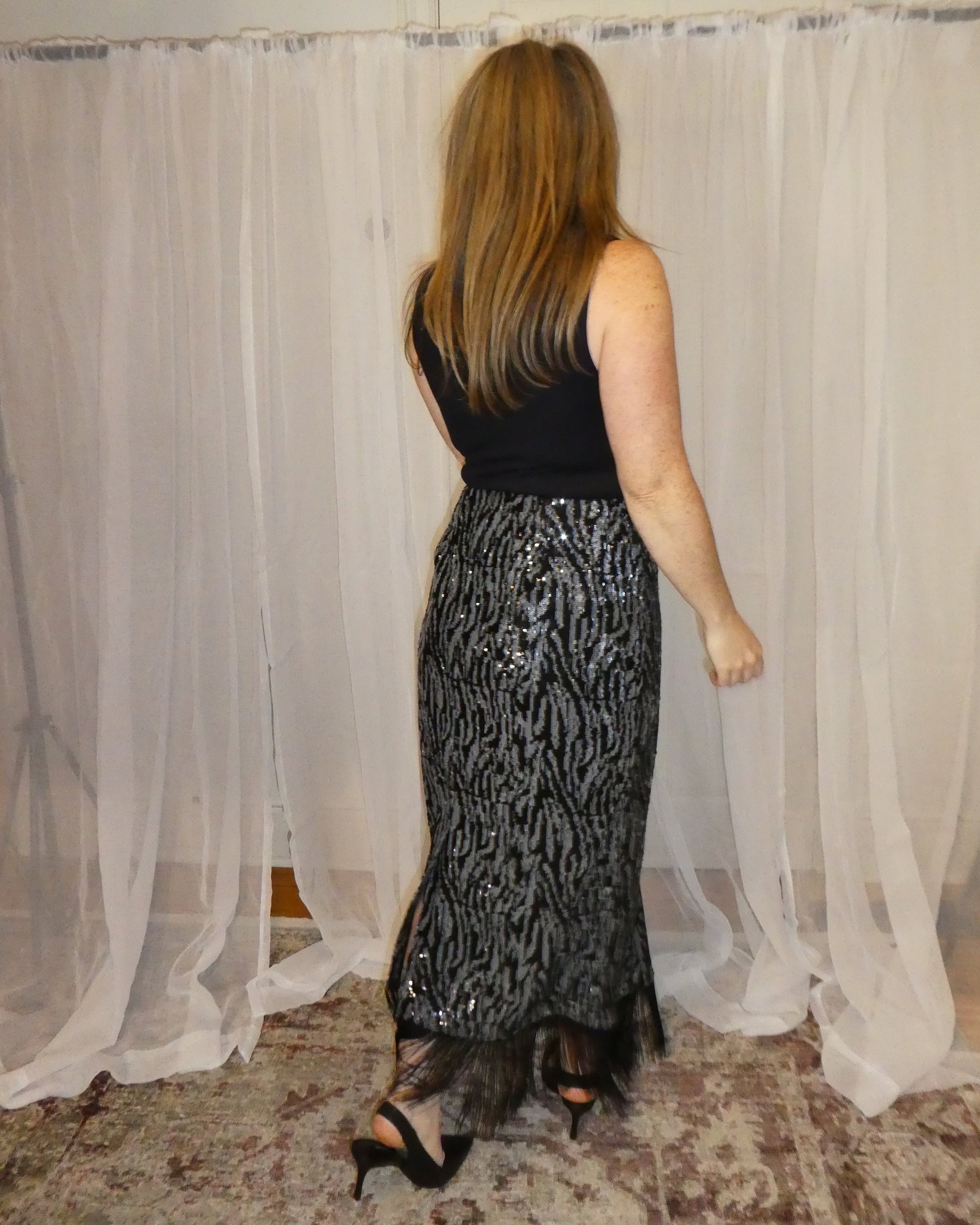 Woman wearing a black top and patterned skirt standing in front of a white curtain.