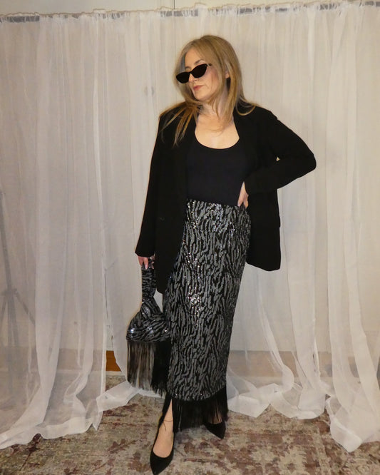 Woman posing in front of a white curtain wearing sunglasses, black top, and patterned skirt.