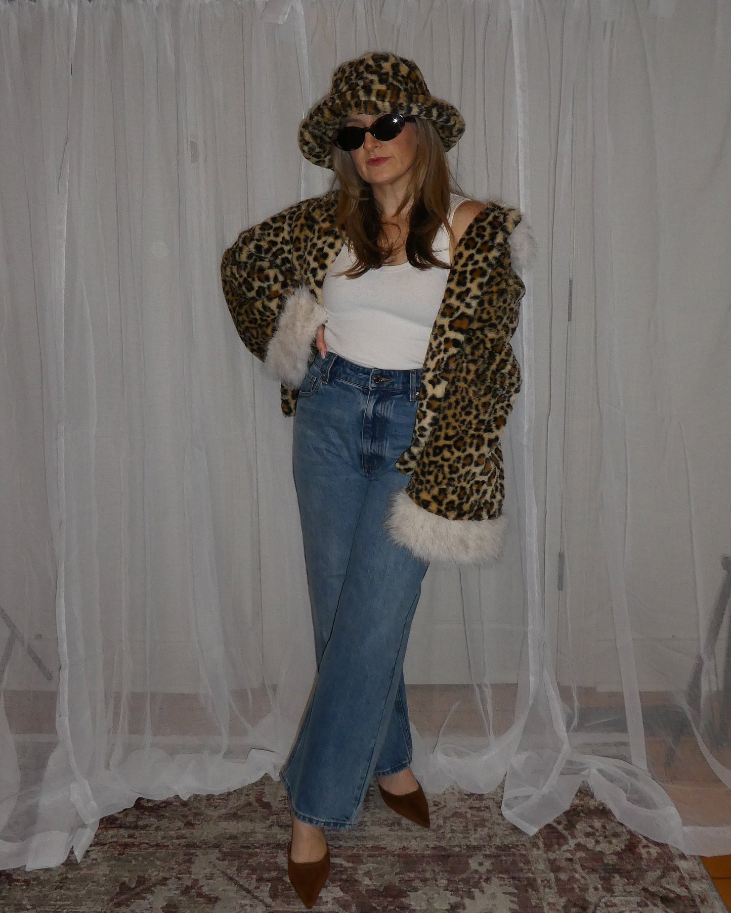 Person wearing a leopard print coat with fur trim, sunglasses, and jeans in front of sheer curtains.